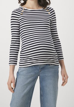 Vrouw draagt een zwart-wit gestreept shirt met lange mouwen en gouden knopen op de schouders, gecombineerd met lichtblauwe spijkerbroek.