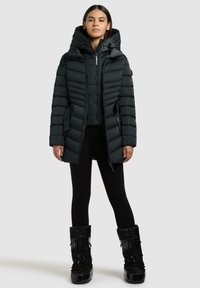 Sort pufferjakke med bred kanalquiltedesign, justerbar hætte og sidelommer, båret over sorte leggings og høje sorte støvler.