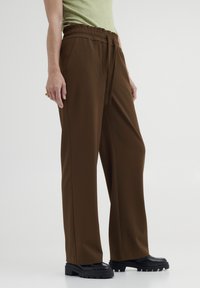 Braune weite Hosen mit einem Kordelzugbund, aus glattem Stoff gefertigt. Kombiniert mit schwarzen Schuhen mit dicker Sohle, die eine strukturierte Sohle aufweisen.