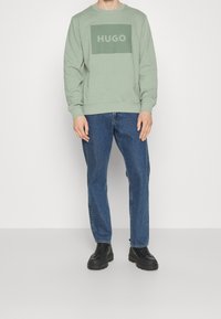Sudadera verde claro con un logo rectangular gris que dice "HUGO" en el centro, combinada con vaqueros azules y zapatos negros con suela gruesa.