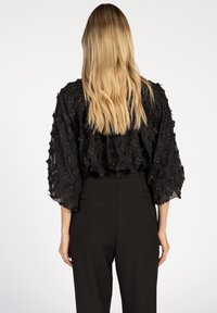 Zwarte blouse met een gestructureerd bloemenpatroon, doorschijnende stof en wijde mouwen, gecombineerd met hooggerokte zwarte broek. Glad haar valt over de schouders.