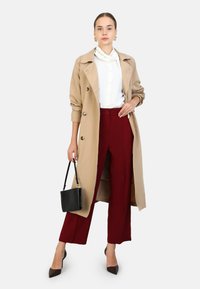 Beige trenchcoat met grote knopen, witte blouse, bordeaux hoge taille broek, zwarte handtas en pumps met spitse neuzen.