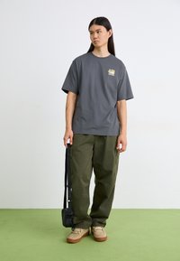 Tricou gri din bumbac, cu mâneci scurte și un grafic pe pieptul stâng, purtat cu pantaloni cargo verzi măslinii și adidași bej. Geantă neagră tip crossbody.