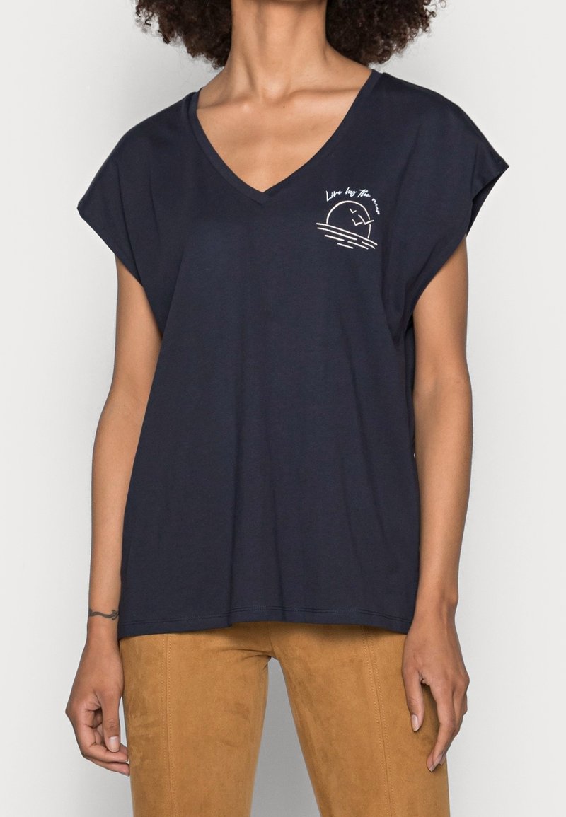 T-shirt noir à col en V avec manches courtes, arborant un petit motif blanc et le texte « Live by the sea » sur le côté gauche de la poitrine, portée avec un pantalon marron.