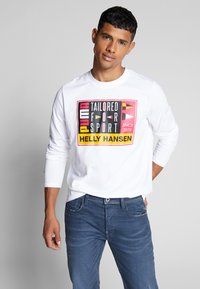 Tricou alb cu mânecă lungă, având un design grafic multicolor cu text, asortat cu blugi albaștri. Gât simplu și croială lejeră.