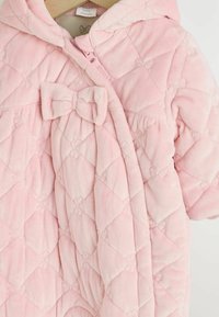 Manteau pour bébé en polaire douce, matelassé et rose, avec un détail de nœud à l'avant, une capuche et une fermeture éclair. Motif en losanges texturé sur l'ensemble.