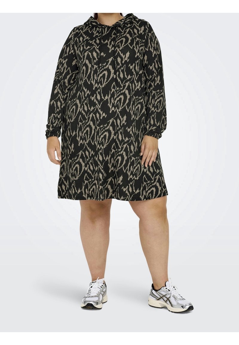 Personne portant une robe à capuche aux motifs abstraits noirs et beiges, associée à des chaussures de sport blanches et noires, debout devant un fond uni.