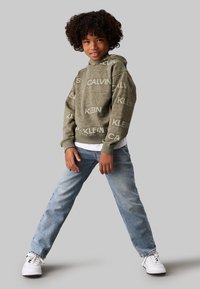 Felpa verde oliva con logo ripetuto "Calvin Klein", abbinata a jeans in denim azzurro chiaro e sneaker bianche. Vestibilità casual, spessore medio.