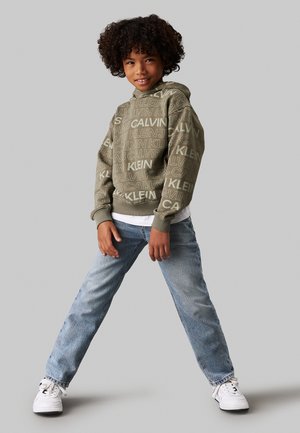 Felpa verde oliva con logo ripetuto "Calvin Klein", abbinata a jeans in denim azzurro chiaro e sneaker bianche. Vestibilità casual, spessore medio.