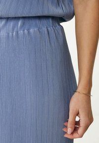 Jasnoniebieski, plisowany materiał w pionowe paski. Posiada elastyczny pas i gładką teksturę. Złota bransoletka na nadgarstku.