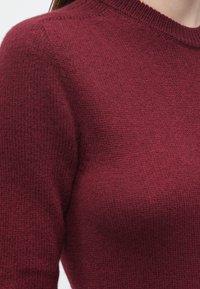 Bordowy sweter z dzianiny z okrągłym dekoltem, krótkimi rękawami i teksturowanym wykończeniem. Zbliżenie ukazuje szczegóły tkaniny i szwów.