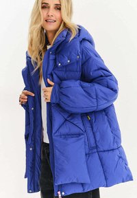 Modrá prošívaná bunda s kapucí, opatřená patentními knoflíky, prošívanou texturou, zipovým zapínáním a několika kapsami. Materiály vypadají jako syntetické.