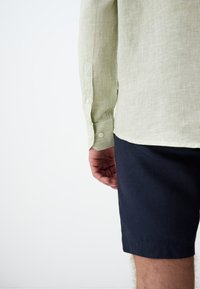 Hellgrün gestreiftes Hemd mit langen Ärmeln, dazu sind eine dunkelblaue Shorts sichtbar. Der Stoff sieht strukturiert und leicht zerknittert aus.