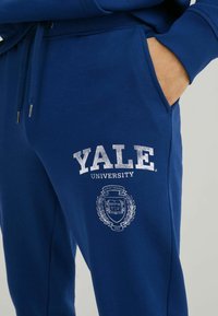 Blue sweatpants made of cotton blend, featuring white "YALE UNIVERSITY" text and emblem with a laurel design on the left thigh.