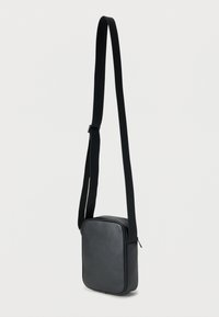 Μαύρη δερμάτινη τσάντα χιαστί με υφή στην επιφάνεια, ορθογώνιο σχήμα και ρυθμιζόμενο υφασμάτινο λουρί. Διαθέτει φερμουάρ κλεισίματος.