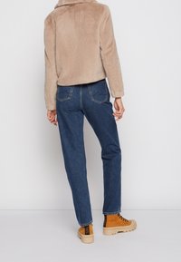 Person som bär en beige fuskpälsjacka, blå högmidjade jeans och beige snörade stövlar, stående mot vit bakgrund.