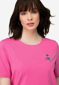 Roze katoenen T-shirt met een ronde hals. Heeft een geprinte ankerafbeelding en tekst aan de linkerkant. Zachte textuur en korte mouwen.