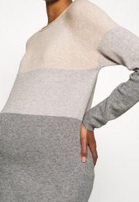 Person som bär en långärmad stickad klänning med beige, ljusgrå och mörkgrå färgblock, med handen vilande på höften.