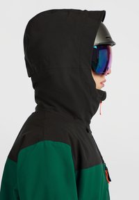 Personne portant une veste noire et verte à capuche, un casque sombre et des lunettes de ski réfléchissantes, regardant vers la droite sur un fond uni.