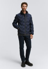 Man som bär en marinblå kviltad jacka med dragkedjefickor, mörka byxor och svarta stövlar, stående mot en slät ljus bakgrund.