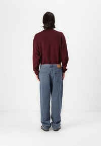 Burgunderfärgad sweatshirt med rund hals och långa ärmar, kombinerad med lösa blå jeans som har två bakfickor och en läderpatch vid midjan.