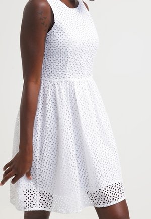 Robe blanche sans manches en tissu ajouré avec corsage ajusté et jupe évasée portée par une personne à la peau foncée avec des tatouages d'oiseaux sur le haut du bras.