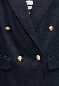 Blazer bleu marine foncé à double boutonnage, avec quatre boutons dorés, associé à un haut rayé blanc et bleu marine visible en dessous.