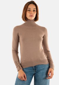 Eine Frau mit schulterlangem Haar, die einen beigen Rollkragenpullover und eine blaue Jeans trägt, steht vor einem einfarbigen Hintergrund.