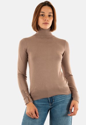 Eine Frau mit schulterlangem Haar, die einen beigen Rollkragenpullover und eine blaue Jeans trägt, steht vor einem einfarbigen Hintergrund.