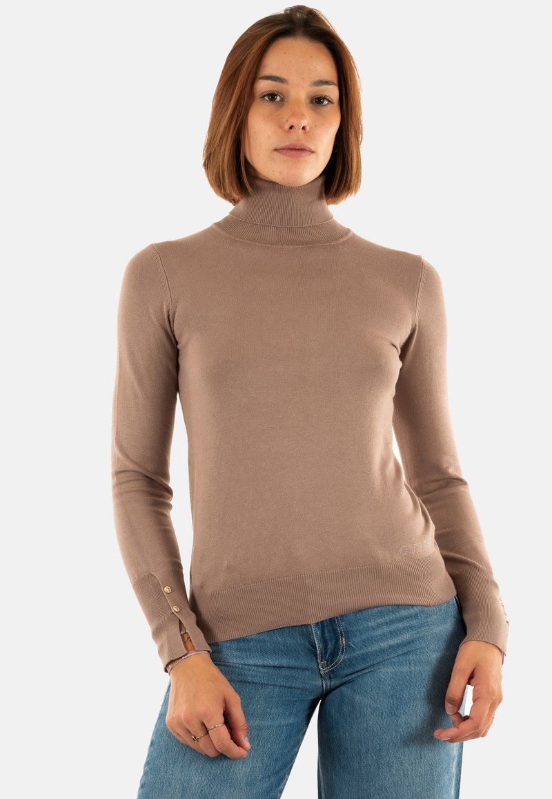 Eine Frau mit schulterlangem Haar, die einen beigen Rollkragenpullover und eine blaue Jeans trägt, steht vor einem einfarbigen Hintergrund.
