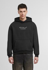 Crna dukserica s kapuljačom napravljena od mekog materijala; ima prednji džep i otisnuti tekst na arapskom i engleskom: "Prvo voli sebe."