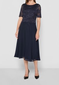 Mörkblå klänning med ett spetsöverdel och korta ärmar, en flödande kjol och en mjuk textur. Svarta spetsiga klackskor kompletterar outfiten.