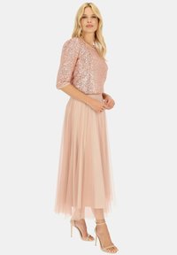 Top z cekinów w kolorze różowego złota z rękawami o długości trzech czwartych, w połączeniu z lekką, różową spódnicą z tiulu. Minimalistyczne beżowe sandały dopełniają całość.