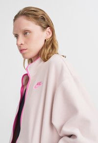 Roza flis jakna s svijetloružičastim patentnim zatvaračem i logom. Ima visoki ovratnik i ležeran kroj. Mekana tekstura, minimalistički dizajn i sportski stil.