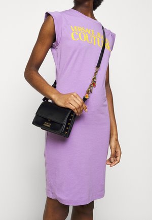 Fioletowa bawełniana sukienka z krótkimi rękawami, z napisem "VERSACE JEANS COUTURE" w kolorze żółtym, zestawiona z czarną skórzaną torebką na ramię zdobioną złotymi ćwiekami.