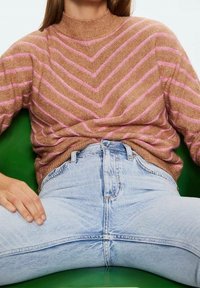 Jersey marrón con rayas en forma de chevron rosa, cuello alto, combinado con vaqueros azul claro de tiro alto, sentado sobre un fondo verde.