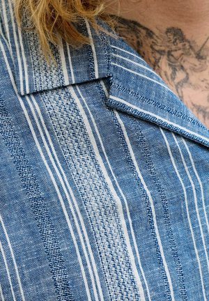 Sinine-valge triibulise kangast krae lähivaade särgil, mille kohal on nähtav osa tätoveeringust ja blondid juuksed.