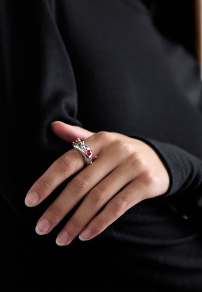 Hand met natuurlijke nagels die een zilveren ring met meerdere kleurrijke edelstenen draagt, rustend tegen een zwart shirt met lange mouwen.