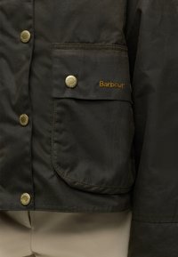 Poche de veste Barbour vert foncé avec bouton doré et logo Barbour brodé en fil jaune sur le rabat de la poche.