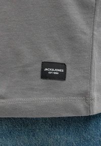 Camiseta de algodón gris con un corte recto, que presenta una etiqueta de tela negra con la inscripción "JACK & JONES EST. 1990" en el dobladillo.
