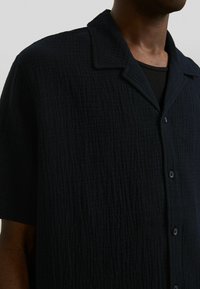 Lyhythihainen, tummansininen paita, joka on valmistettu teksturoidusta kankaasta, jossa on hienovarainen rypytetty kuvio; siinä on lovikaulus ja napitus etuosassa.