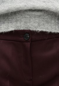 Κοντινό πλάνο γκρι πλεκτού πουλόβερ που είναι βαλμένο μέσα σε παντελόνι με ψηλή μέση σε μπορντό χρώμα με μαύρο κουμπί κλεισίματος.