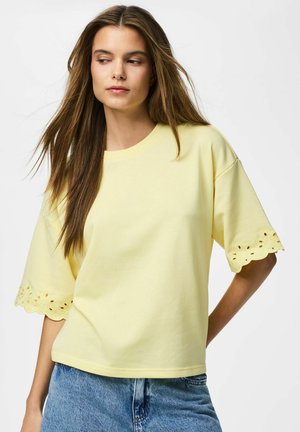 Žena s dlhými hnedými vlasmi, oblečená v voľnom bledom žltom top-e so zvlnenými dierkovanými rukávmi a modrých rifliach, pozerá sa do strany.