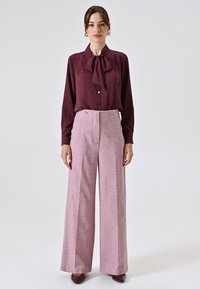 Blusa bordeaux con dettaglio al collo a fiocco, abbinata a pantaloni ampi a vita alta di colore rosa, caratterizzati da una sottile texture e due bottoni frontali.