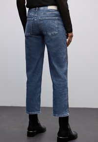 Jeans de mezclilla azul con un diseño de pierna recta y cintura media, y con bolsillos traseros. Presenta una franja lateral texturizada. Combinados con botas negras hasta el tobillo.