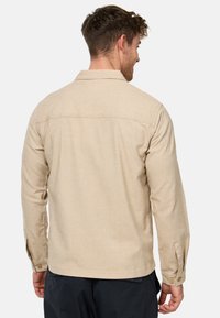 Chemise beige à manches longues, avec des poignets boutonnés et un col. Elle présente une texture lisse et un design simple et épuré.