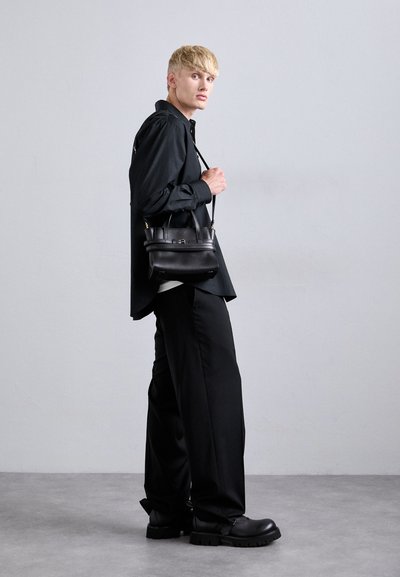 O tânăr cu păr blond stă deoparte, îmbrăcat cu pantaloni negri largi, un tricou negru, cizme negre masive și poartă o geantă mică și neagră pe umăr.