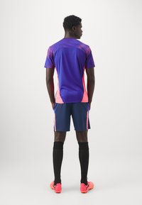 Camisola atlética roxa e rosa com parte superior texturizada, combinada com calções azul-marinho. Meias altas pretas até ao joelho e sapatilhas brilhantes rosas completam o visual.