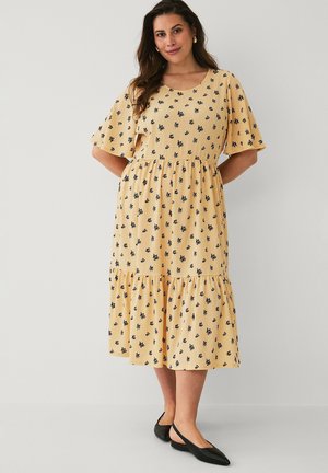 Kvinde iført en gul midikjole med sort blomstermønster, korte flagrende ærmer, smøg i taljen og sorte spidse slingback-flats.