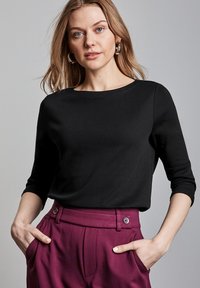Schwarzes Oberteil mit drei Viertel Ärmeln aus glattem Stoff, mit rundem Halsausschnitt. Kombiniert mit hoch taillierten, pflaumenfarbigen Hosen mit Knopfdetails.